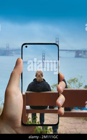 Mit einem Mobiltelefon ein Foto von einer Frau auf einer Bank machen, die die Golden Gate Bridge in San Francisco, USA, beobachtet. Stockfoto