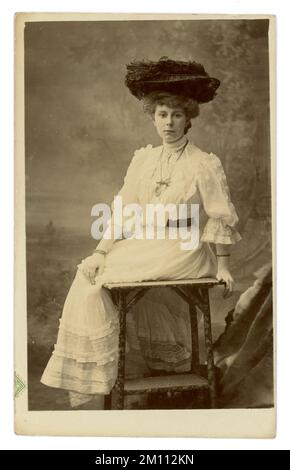 Die ursprüngliche Postkarte aus der Edwardianischen Ära war eine schöne edwardianische Frau, die einen großen, aufwendigen Hut mit Straußenfedern trug, ein weißes Sommerkleid, fließende Ärmel, Handschuhe, eine für die Möbel dieser Zeit typische Bambus-Tischstütze trug. Edwardianerin. Edwardianerin. Datiert/veröffentlicht am 20. November 1906, Sheffield, England, Großbritannien Stockfoto