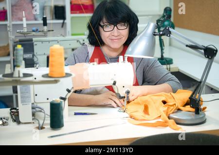 Eine hübsche lächelnde Frau mit Brille sitzt an einer Nähmaschine Stockfoto