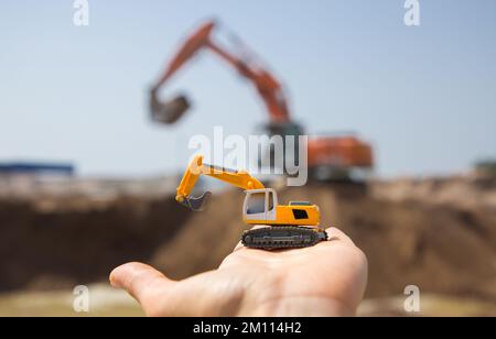 Auf dem Arm befindet sich ein kleiner orangefarbener Spielzeugbagger. Hinter ihm, außer Fokus, ist ein echter arbeitend Bagger in einem Steinbruch. Ein heller, sonniger Tag. Betrieb der Konstr Stockfoto
