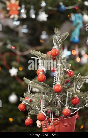 Eine vertikale Aufnahme einer kleinen Tanne mit Dekorationen in einem roten Eimer mit dem Weihnachtsbaum im Hintergrund Stockfoto
