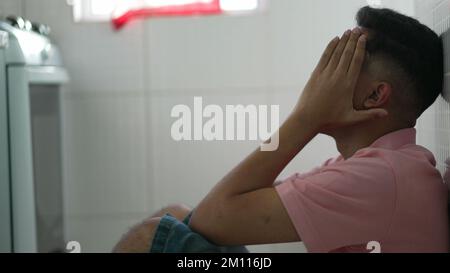 Ängstlicher junger südamerikanischer Mann, der auf dem Boden sitzt und sich besorgt fühlt. Gestresste Person, die sich von modernen Problemen überwältigt fühlt. Einsamer brasilianischer gu Stockfoto