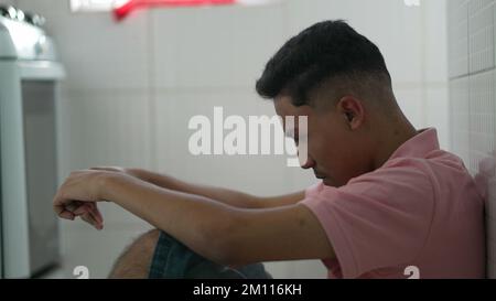 Ängstlicher junger südamerikanischer Mann, der auf dem Boden sitzt und sich besorgt fühlt. Gestresste Person, die sich von modernen Problemen überwältigt fühlt. Einsamer brasilianischer gu Stockfoto