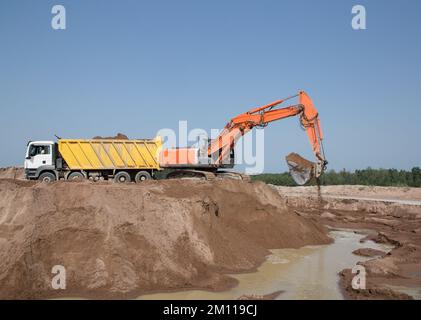 Die große Schaufel eines orangefarbenen Raupenbaggers gießt Erde in einen gelben Kipplaster. Arbeitszeitpunkt auf einer Baustelle an einem sonnigen Tag gegen ein Blau Stockfoto