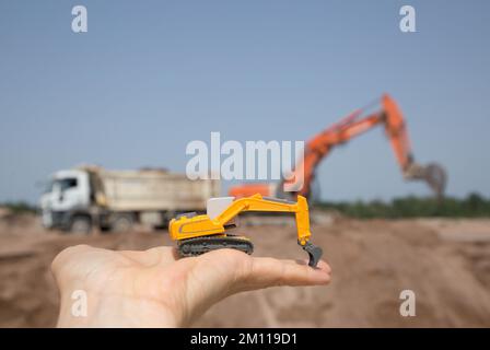 Am Arm befindet sich ein kleiner orangefarbener Spielzeugbagger. Hinter ihm, unscharf, ist ein echter Bagger und Kipplaster, der in einem Steinbruch arbeitet. Funktionsweise der Kontroll Stockfoto