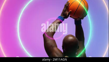 Ein afroamerikanischer Basketballspieler mit Glatze wirft den Ball über beleuchtete mehrfarbige Kreise. Composite, Kopierbereich, Sport, Wettkampf, Illustration, Stockfoto