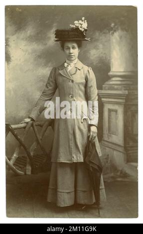 Original Edwardians Atelierpostkarte mit attraktiver Frau in langer Jacke und Rock, großem Hut mit Rüschenspitze und Blumenzweig, mit einem Regenschirm mit Stock Griff. Edwardianerin. Edwardianerin. Juli 1910, Großbritannien Stockfoto