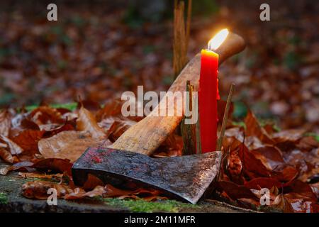 Das Weihnachtskartenmotiv für Holzfäller. Eine brennende Kerze mit einer Axt zwischen verwelkten Blättern auf einem Baumstumpf im Wald. Stockfoto