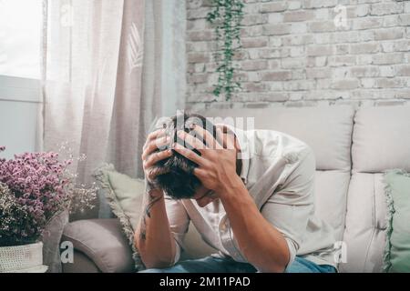 Ein junger kranker Mann, der auf dem Sofa sitzt und sich schlecht fühlt, mit Schmerzen und Kopfschmerzen. Ein Teenager mit einer Tätowierung am Arm, der sein Gesicht berührt, ist krank. Stockfoto