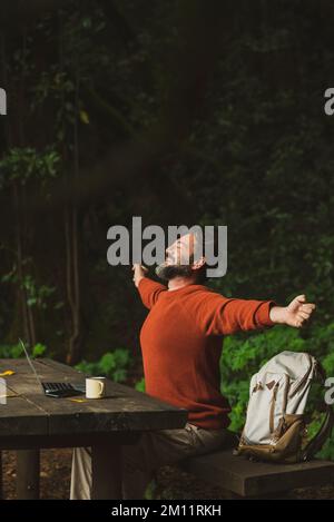 Ein Reisender Mann streckt sich nach der Arbeit die Arme im Naturwald aus, mit Laptop und Internet-Verbindung. Das Konzept des Menschen und der digitale Lebensjob. Glücklicher Erwachsener mit Rucksack. Grün Stockfoto