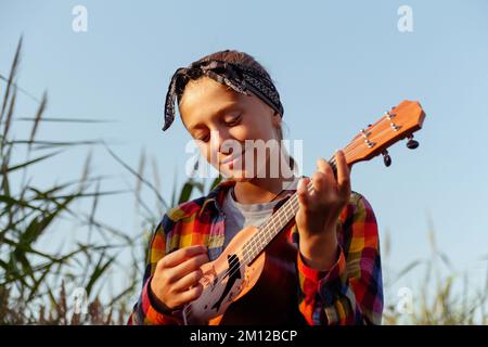 Unschärfe-Mädchen mit Gitarre. Ein Mädchen, das in der Natur herumläuft. Ein kleines Mädchen draußen auf der grünen Wiese. Ukulele-Akkord. Sommerzeit. Ein Volkskünstler Stockfoto