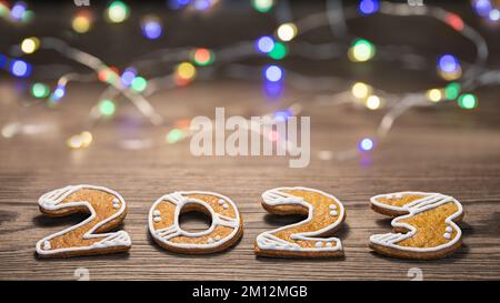 Wunderschöne Neujahrskarte für Happy 2023 aus kunstvoll verzierten Lebkuchenzahlen und LED-Lichterketten. Nahaufnahme von süßen Plätzchen mit Weihnachtsgebäck auf Holz. Bokeh. Stockfoto