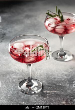 Zwei Gläser Granatapfelcocktail mit Rosmarin auf grauem, strukturiertem Hintergrund, isoliert aus nächster Nähe. Weihnachten oder Valentinsgetränk. Vertikale Ausrichtung Stockfoto