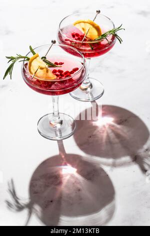 Zwei Weine aus Granatapfel mit Rosmarin und Zitrone auf weißem Marmorhintergrund, isoliert mit klaren Schatten in rauem Licht. Weihnachten oder Vale Stockfoto