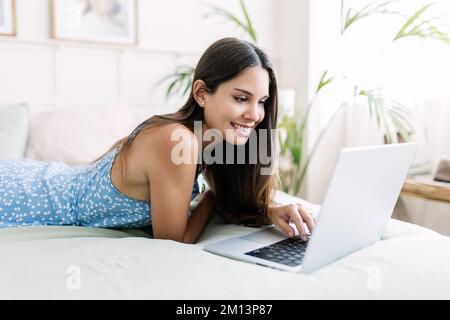 Fröhlich entspannte junge Erwachsene Frau, die zu Hause auf dem Bett liegend ein Notebook benutzt Stockfoto