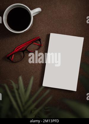 Flache Ansicht von oben mit weißer, weicher Cover Book Mockup, Kaffeebecher, Gläser und verschwommener Pflanze im Vordergrund auf einem braunen Holztisch. Stockfoto