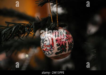 Nahaufnahme einer bemalten Weihnachtskugel, dekoriert mit fröhlichen Poinsettias und Cartoons, die auf einem Ast mit warmgelbem Hintergrund hängen Stockfoto