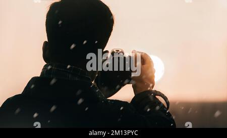 Eine Nahaufnahme eines Mannes, der eine Kamera in der Hand hält und ein Foto von der Sonne bei Sonnenuntergang macht, umgeben von Schneeflocken Stockfoto