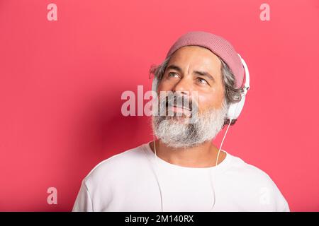 Ein weißer, fröhlicher Mann mit grauhaarigem Bart in lässigem pinkfarbenen Hut und Pullover, der Musik mit Kopfhörern hört, die auf dem Hintergrund einer fuchsianischen Wand isoliert sind Stockfoto