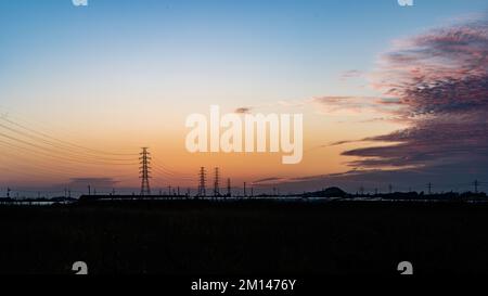 Ein dramatisches Landschaftsfoto bei Sonnenuntergang über einer weiten ländlichen Ebene mit Stromleitungen und Strommasten, die in der Abenddämmerung in den fernen Horizont zurückziehen Stockfoto