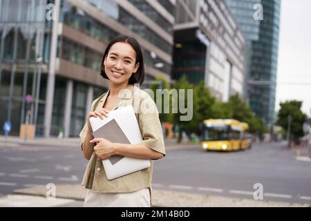 Schöne asiatische Mädchen lächelt als pendelt zur Arbeit, steht auf der Straße mit Laptop und Notebook Stockfoto