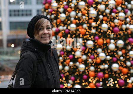 Die beste Weihnachtszeit für das ältere Modell am großen Weihnachtsbaum mit einem Lächeln im Gesicht. Frau im besten Alter steht vor einem großen Weihnachtsbaum glücklich Stockfoto