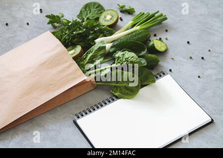 Einkaufstasche aus umweltfreundlichem Papier mit frischem grünen Gemüse, Kräutern, Obst neben einem leeren Notizblock auf dem Küchentisch Stockfoto