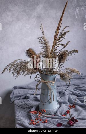 Getrocknete Pflanzen und Blumen in einer Vintage-Vase mit einem kleinen Herz auf einem Tisch mit einem Schal darauf Stockfoto