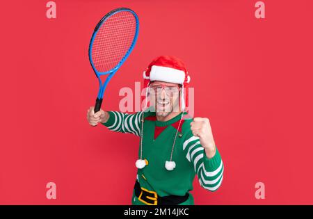 Glücklicher erfolgreicher Mann im Elfenkostüm und weihnachtsmann-Hut. der weihnachtsmann hält eine Tennisrakete Stockfoto