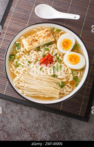 Asiatische Ramen-Suppe mit Nudeln, Ei, Tofu und Enoki aus nächster Nähe in einer Schüssel auf dem Tisch. Vertikale Draufsicht von oben Stockfoto