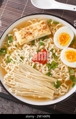 Asiatische Suppe mit Tofu, Enoki-Pilzen, Nudeln, grünen Zwiebeln und Chili-Paprika in einer Schüssel auf dem Tisch. Vertikale Draufsicht von oben Stockfoto