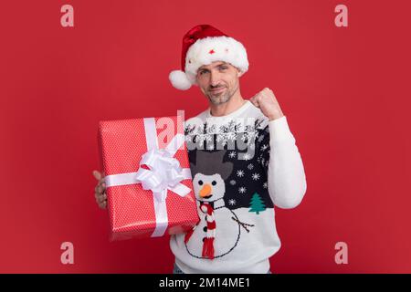 Weihnachtsmann mit Gewinnerschild, Geschenk auf rotem Studiohintergrund, glücklicher junger schwarzer Mann mit Weihnachtsmannmütze und Pullover zu Weihnachten oder Neujahr Stockfoto