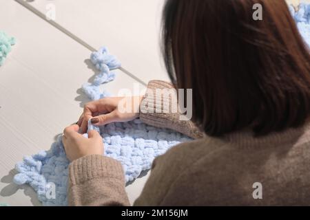 Garn, das mit den Fingern in ein Muster bearbeitet wird. Keine Stricknadeln. Rückansicht einer unbekannten Frau mit flauschigem Faden. Blaue Neugeborenen-Decke ist da Stockfoto