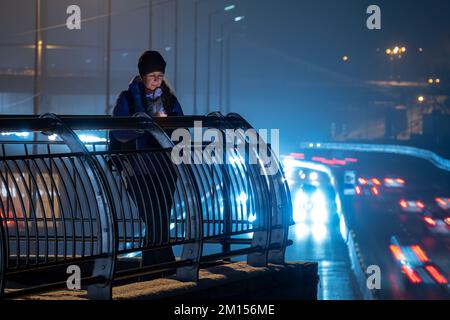 Eine Frau auf der Nachtstraße sieht in ihrem Smartphone mit Autoleuchten im Hintergrund Stockfoto