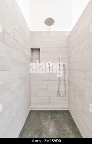 Eine begehbare Dusche mit grauem Fliesenboden und weißen Wandfliesen an den Wänden, es gibt einen Duschkopf über Kopf Stockfoto