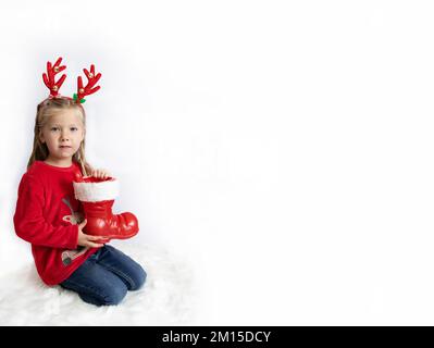 Weihnachten. Ein Mädchen mit Hirschhörnern in einem roten Pullover hält weihnachtsmannstiefel. Banner Stockfoto