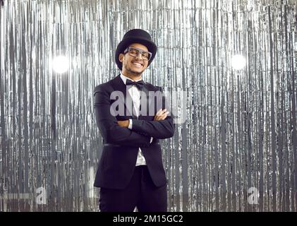 Ein cooler junger, positiver afroamerikanischer Mann im klassischen, eleganten schwarzen Anzug auf silbernem, glänzendem Hintergrund. Stockfoto