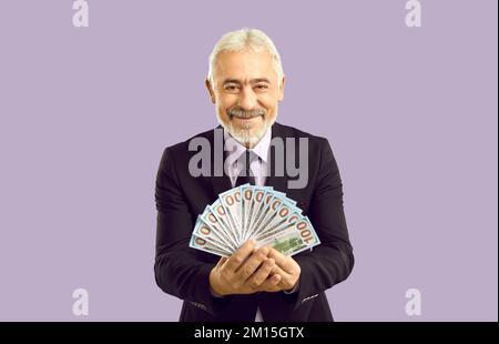 Glücklicher, reifer Mann in formeller Kleidung, der Dollarscheine isoliert auf pastelllila Hintergrund hält. Stockfoto