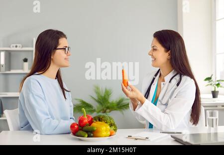 Junge weiße Frau sitzt am Tisch mit Gemüse in der Nähe des Ernährungswissenschaftlers im weißen Kittel Stockfoto