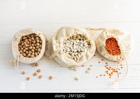 Offene Baumwollsäcke mit organischen Körnern: Linsen, Kichererbsen und Bohnen auf weißem Holzküchentisch. Draufsicht Stockfoto