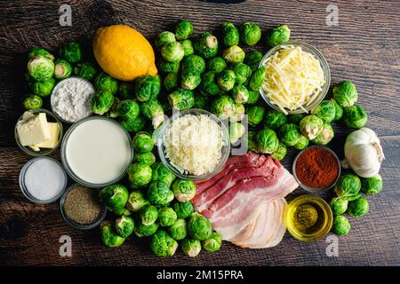 Rosenkohl Gratin mit Speckbestandteilen auf einem Holztisch: Blick von oben auf frische Rosenkohl, Speck, Käse und andere Rohstoffe Stockfoto