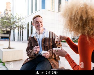 Fröhlicher junger Freund und Freundin, die auf einer Bank an der Stadtstraße sitzen und zusammen lachen, während sie Eiscreme aus Plastikbechern essen Stockfoto