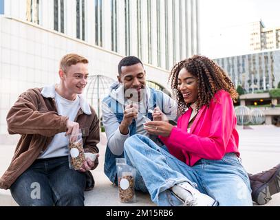 Gruppe multiethnischer Studenten in lässiger Kleidung, die in der Nähe des Universitätsgebäudes sitzen und leckere Snacks essen, während sie sich unterhalten Stockfoto