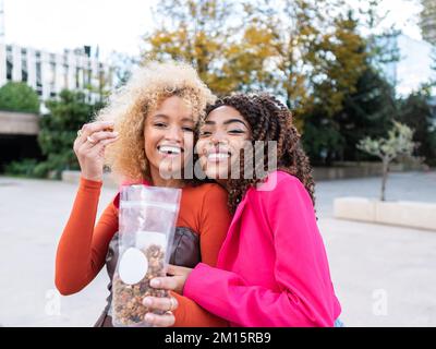 Fröhliche multirassische Freundinnen mit lockigem Haar, die sich auf der Straße umarmen und Nüsse aus Plastikpackungen auf der Straße essen, mit verschwommenem Hintergrund Stockfoto