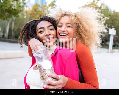 Fröhliche multirassische Freundinnen mit lockigem Haar, die sich auf der Straße umarmen und Nüsse aus Plastikpackungen auf der Straße essen, mit verschwommenem Hintergrund Stockfoto