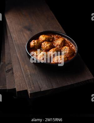 Von oben eine schwarze Schüssel mit köstlichen Fleischbällchen in Tomatensoße und Kräutern, die auf einem Haufen Holzbretter in einem dunklen Raum platziert werden Stockfoto