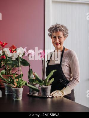 Glückliche reife Frau in Schürze und Handschuhen, die sich um Topforchideen kümmert, während sie in der Küche zu Hause Gartenarbeit macht und in die Kamera schaut Stockfoto