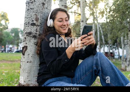 Positive Frau in Kopfhörern, die im grünen Stadtpark mit einem Handy Musik hört, während sie an einem Baumstamm sitzt Stockfoto