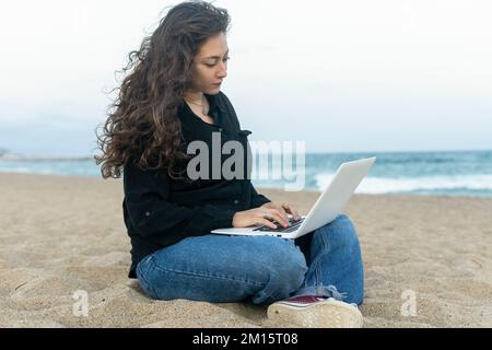 Fokussierte junge Freiberuflerin in lässiger Kleidung, sitzt am Sandstrand nahe dem Meer und arbeitet Remote auf einem Laptop Stockfoto