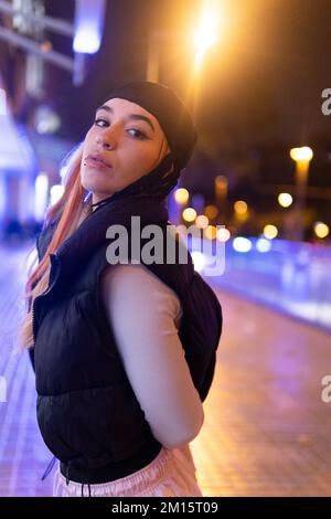 Seitenansicht einer jungen Frau in trendigen Outfits, die sich die Hand hinter dem Rücken hält und über die Schulter in die Kamera schaut, während sie abends auf der Straße der Stadt verbringt Stockfoto